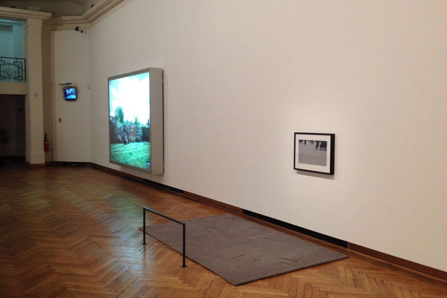 Moderne Kunstgalerie mit Landschaftsgemälde auf einer großen Leinwand links, einem kleineren Schwarz-Weiß-Bild rechts und einem Fernsehbildschirm im Hintergrund, auf Parkettboden.