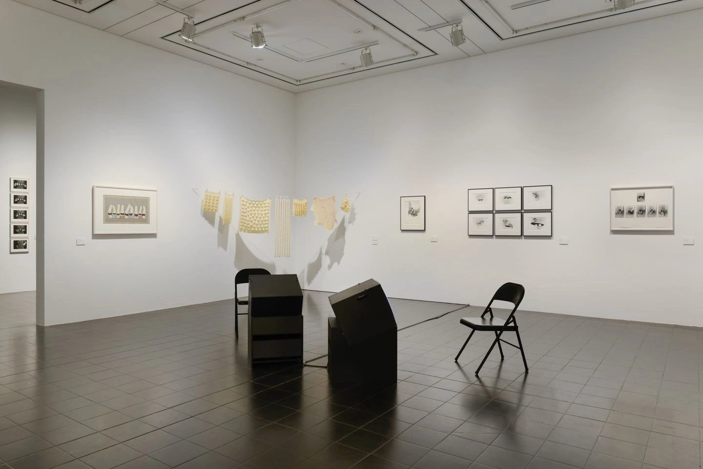 In a somewhat darkened room with white walls and a dark floor, black armchairs stand in front of two black monitors. Various works of art hang framed on the white walls. On the far left, a passage to another room can be seen.