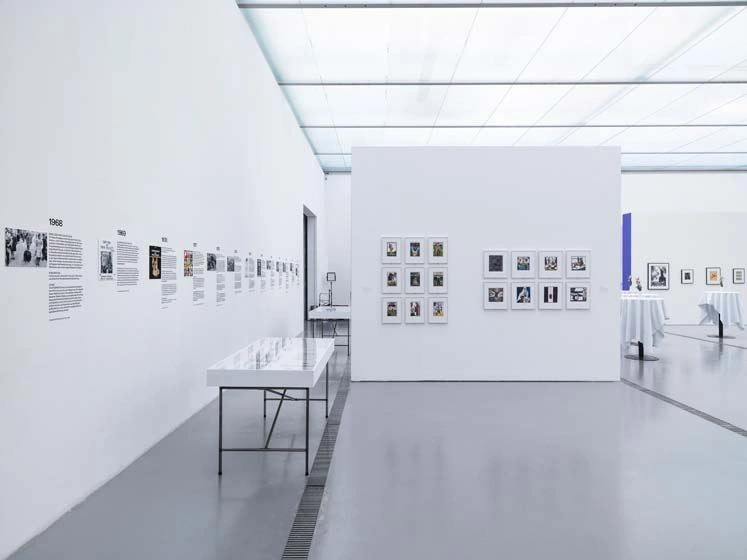 A bright exhibition room with a light gray floor and several photographs on the wall. To the left of the room is a display table with black table legs.