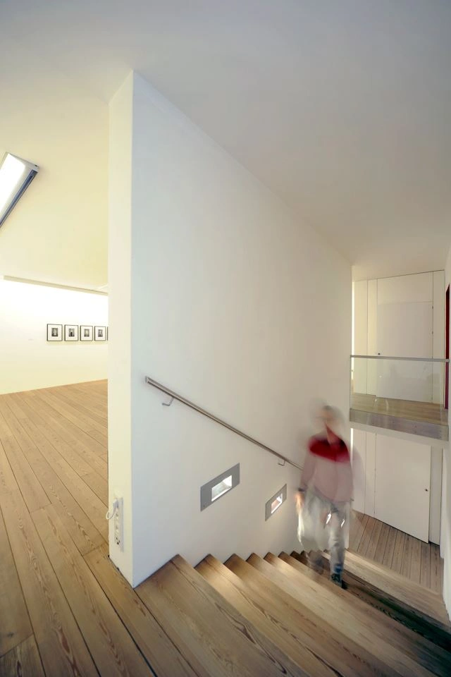 Bright staircase with wooden stairs and white wall. An exhibition visitor can be seen, distorted, climbing the stairs.