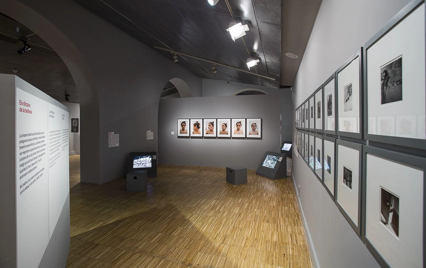 View of a photography exhibition in a room with a vaulted ceiling and wooden floor, featuring numerous framed black-and-white and color photographs on the walls, with screens on stands in the middle of the room and informational text panels at the entrance.