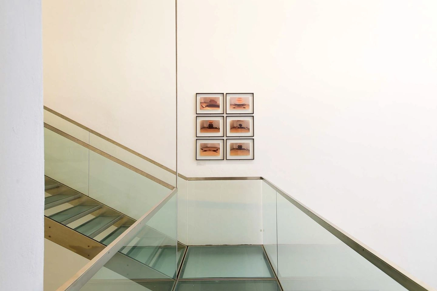 Moderne Museumstreppe aus Glas mit Edelstahlhandlauf und sechs gerahmten Bildern an einer weißen Wand in einer minimalistischen Architekturumgebung.