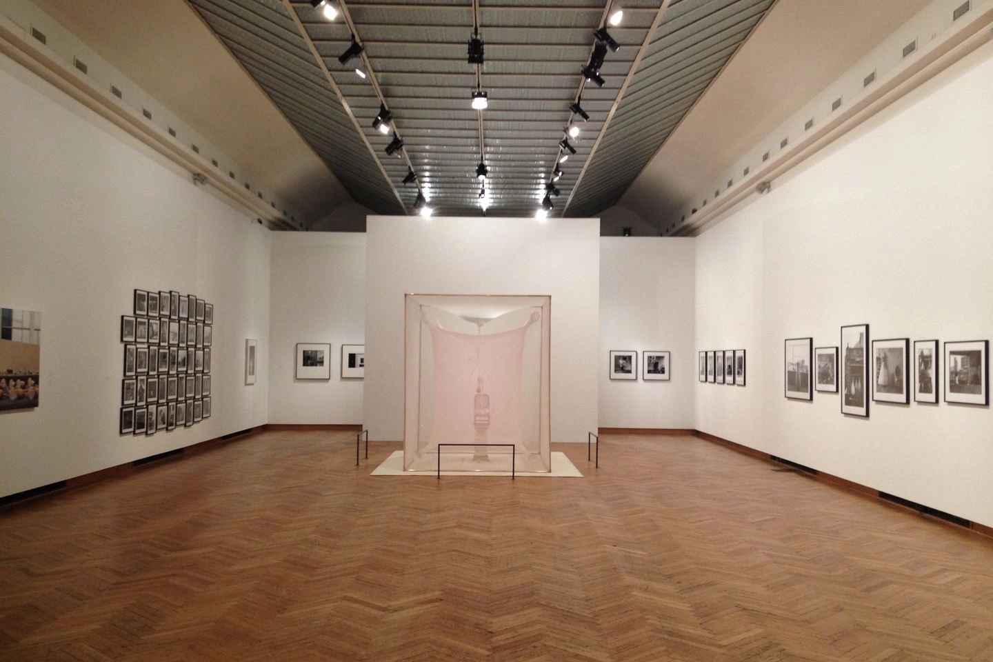 Minimalist exhibition space design in an art hall featuring a central display case containing a sculpture, surrounded by various framed works on the walls and modern lighting under a high ceiling on light parquet flooring.