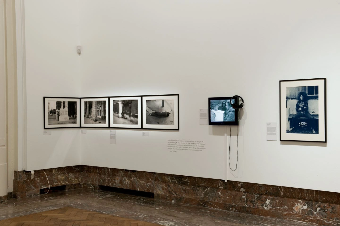 Several works of art by Bozar are displayed in an exhibition hall. On the left are four black and white photographs in landscape format, in the middle a monitor with video and on the right a photograph of a seated person in portrait format.