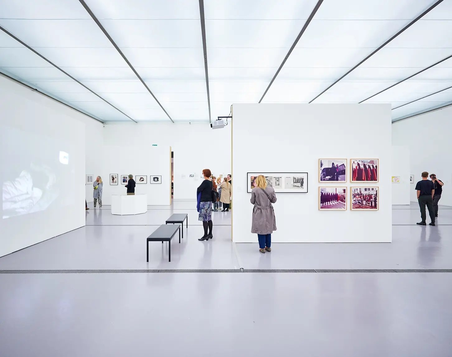 We see the space of an art exhibition showing works of art, painting, photography and moving images. A few interested people are looking at the works of art.