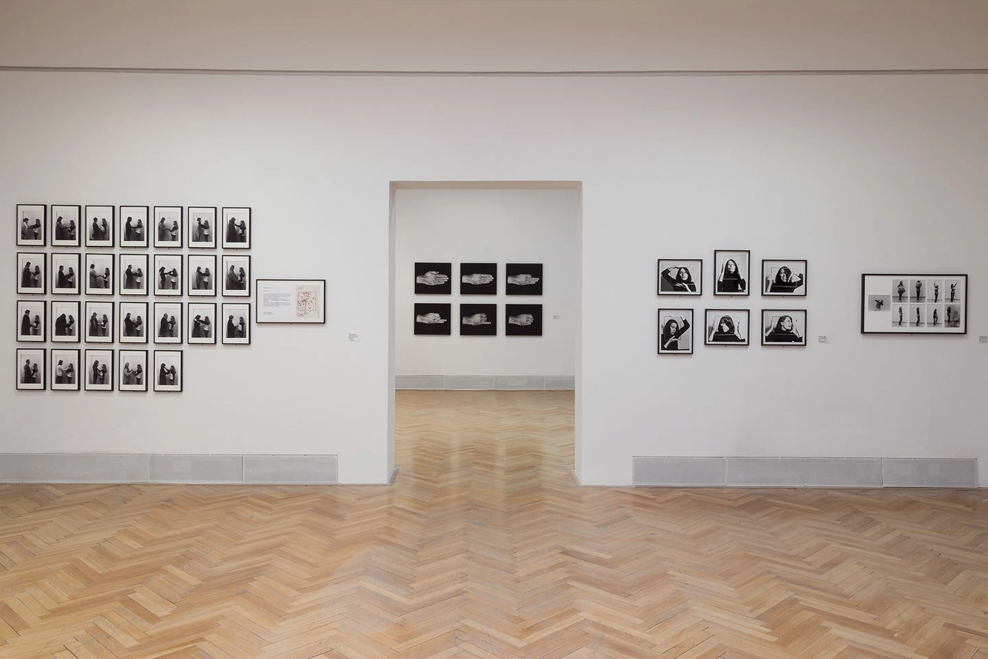 Eine Ausstellungswand in weiß mit Fotographien darauf ist zu sehen. In der Mitte der Wand ist ein Durchgang in einen anderen Ausstellungsraum in dem ebenfalls Fotographien an der Wand hängen.