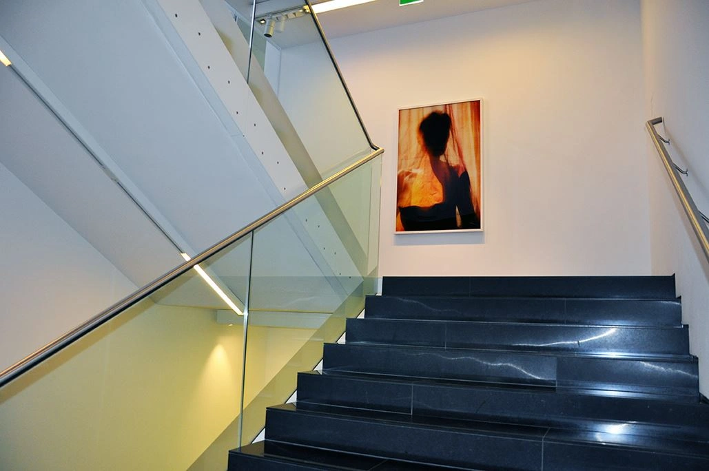 Modern staircase with black steps and glass railing inside a building, featuring an abstract painting on the white wall in the stairwell.