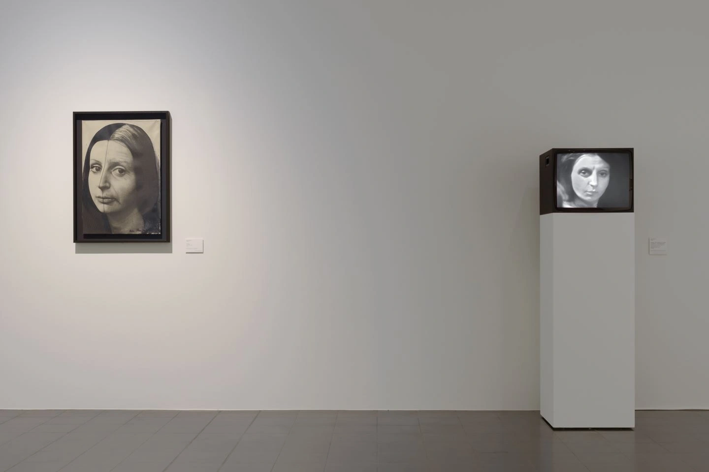 In a somewhat darkened room, a black-and-white portrait photograph of a woman can be seen on a white wall on the left side of the picture. On the right, the same image is displayed on a video monitor.