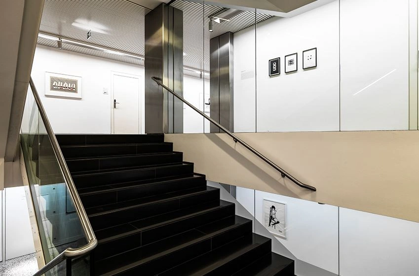 We look into the vertical gallery in the courtyard. In the center is a dark staircase. Works of art from the “Made in Austria” exhibition hang on the white walls.