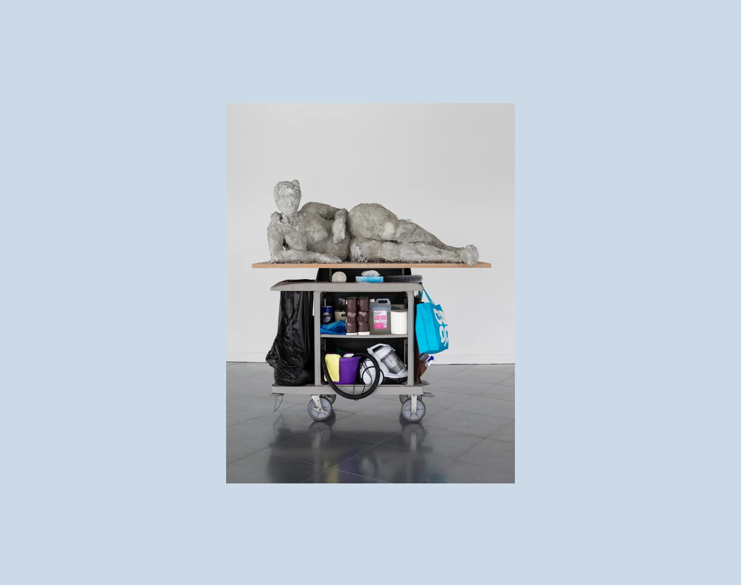 Wir blicken auf ein Kunstwerk von Nicole Wermers. Auf einem Reinigungswagen ist ein Holzbrett angebracht auf dem sich eine steinerne weibliche Figur zu sehen.