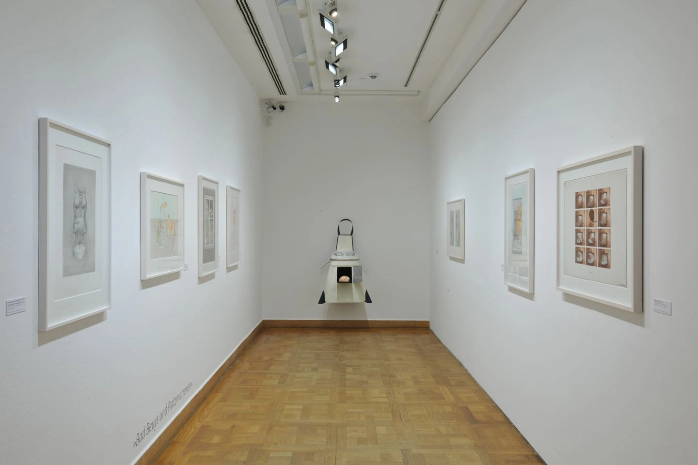 Interior view of an art gallery with white walls featuring several framed black-and-white drawings. A bench is situated in the center of the room, spotlights installed above illuminate the artwork.