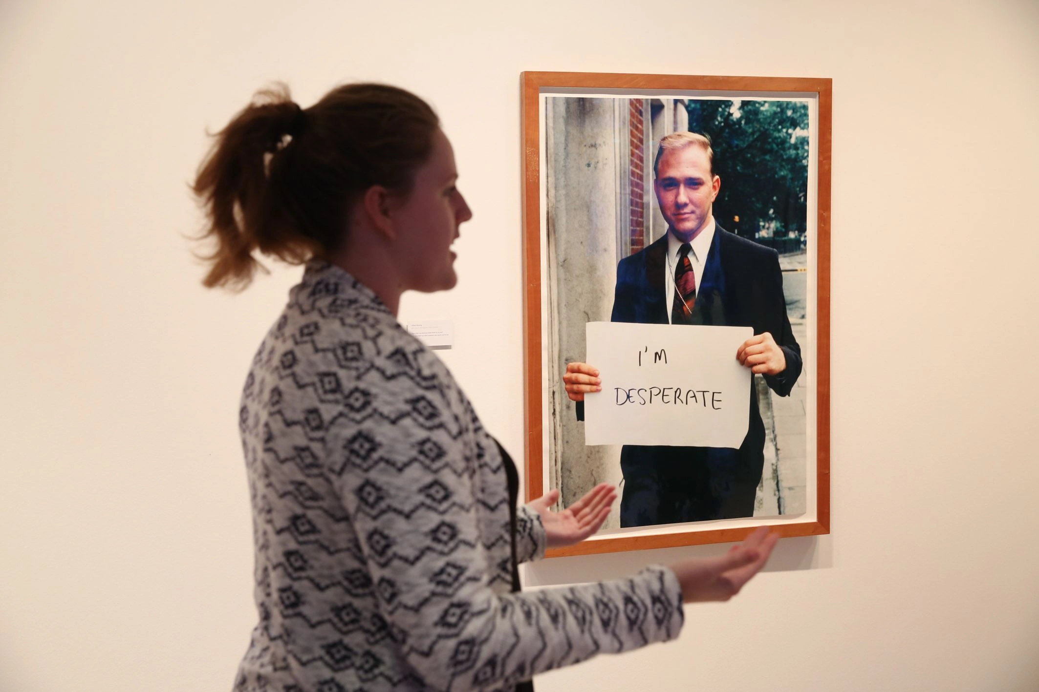 A woman stands in front of a photograph and explains something to an invisible audience. The photograph in the background shows a man with a sign that says I'm Desperate.