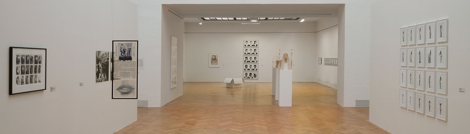 A bright exhibition room with a wooden floor can be seen. Photographs in black and white can be seen on the left and right, and several portrait photographs also hang on the back wall. In the middle of the room are showcases and a white box.