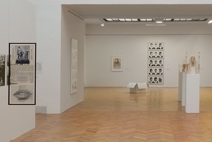 A bright exhibition room with a wooden floor can be seen. Photographs in black and white can be seen on the left and right, and several portrait photographs also hang on the back wall. In the middle of the room are showcases and a white box.