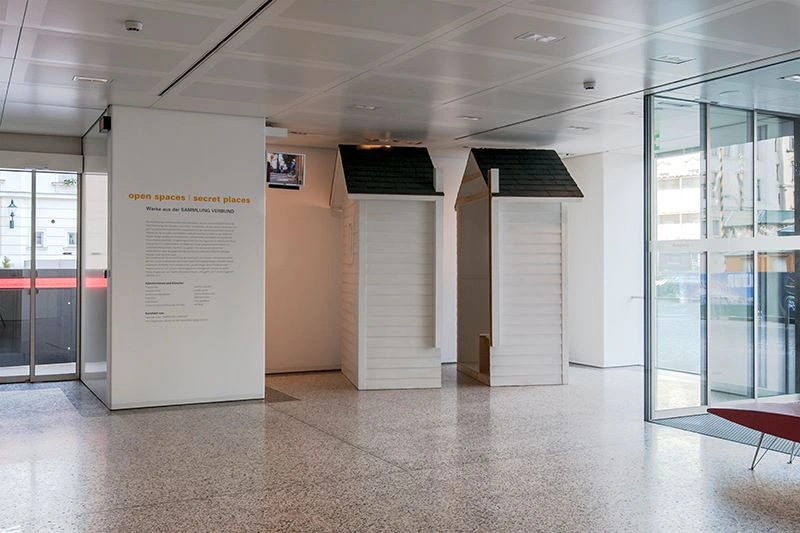 Bright entrance area with two wooden houses with a dark roof. Next to it on a white wall is a text explaining the art installation