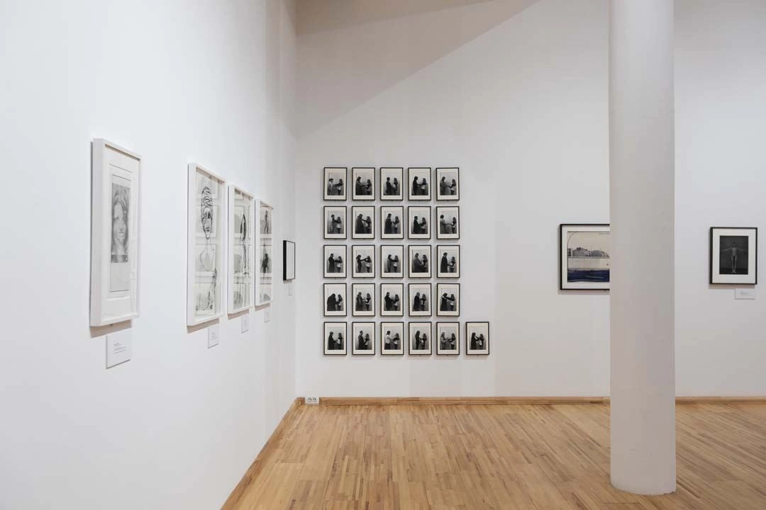 Interior view of a gallery displaying black-and-white photographs arranged in a square composition on a main wall and individual framed images on adjacent walls, over parquet wooden flooring.