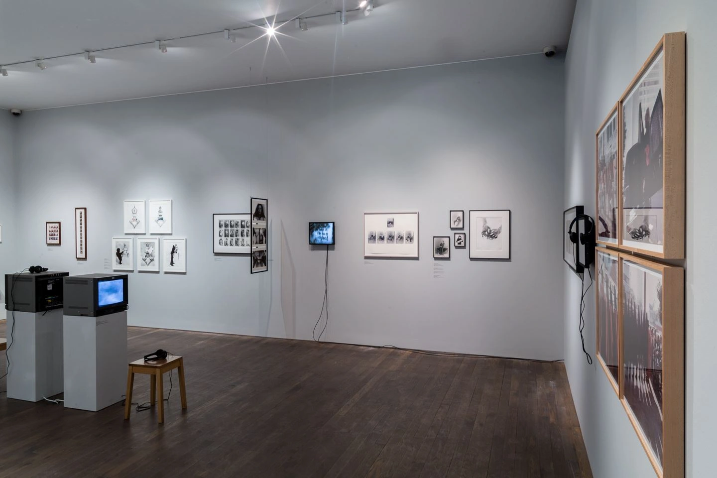 Interior of a contemporary art gallery with a diverse exhibition of framed artworks and photographs on white walls, complemented by audio-visual installations with televisions and headphones on a dark brown wooden floor.