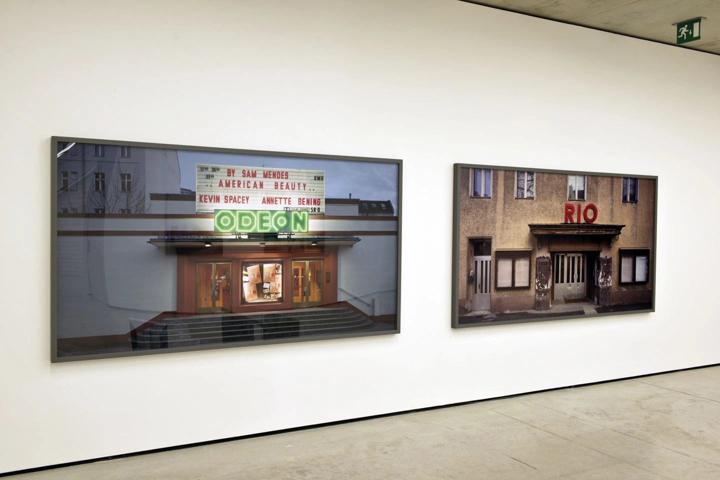 Art gallery displaying two large framed photographs on a white wall, featuring vintage cinema facades with the names "ODEON" on the left and "RIO" on the right side.