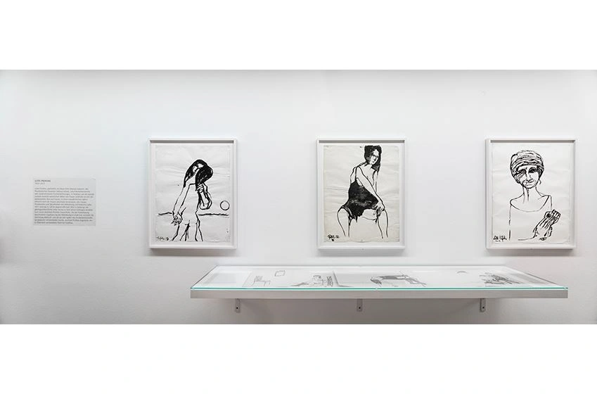 Three ink drawings, each showing women, are at the center of the photograph. Other drawings are also exhibited in a display case below.