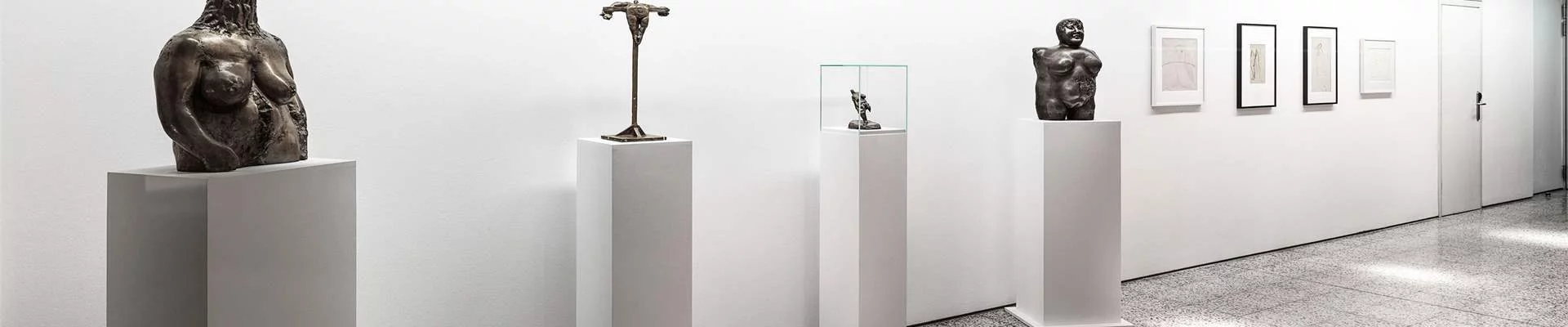 Ein moderner Bürogang mit mehreren abstrakten Skulpturen auf weißen Sockeln ist zu sehen. Rechts daneben hängen hochformatige Bilder.