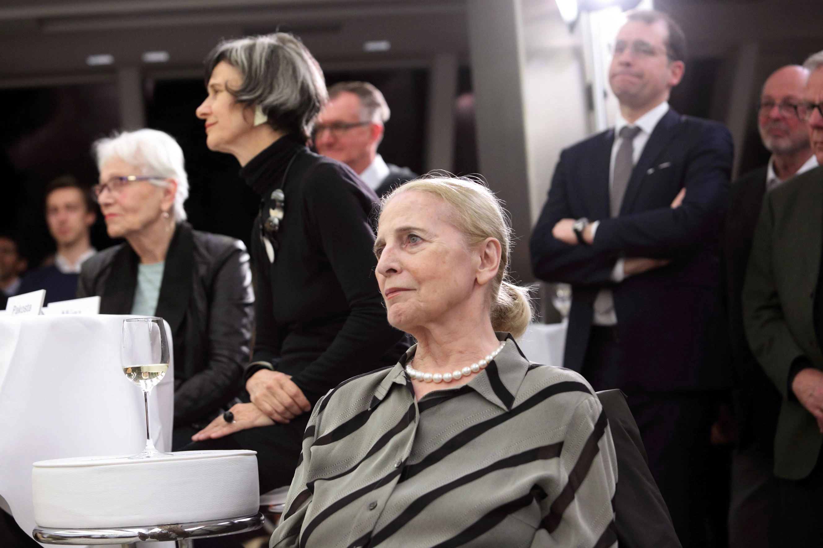 Bei der Pressekonferenz der Ausstellungseröffnung waren zahlreiche Interessierte anwesend. Im Fokus sitzt eine ältere Dame mit blonden Haaren.