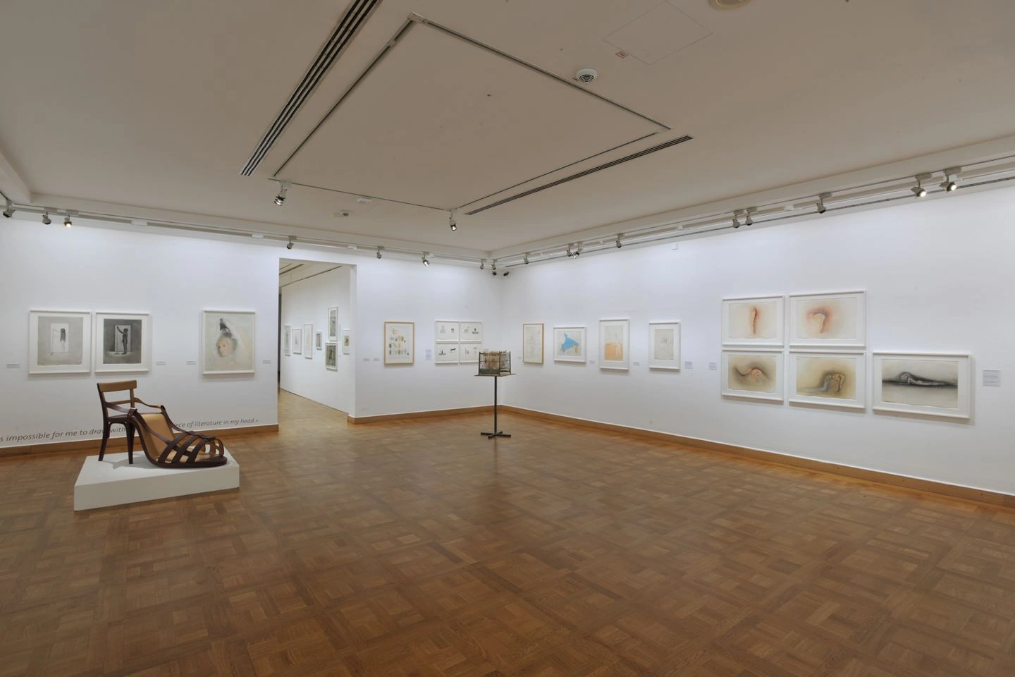 Interior view of a gallery with parquet flooring and white walls, on which various framed artworks are hung, including photographs and drawings. A modern wooden lounger is on the left side.