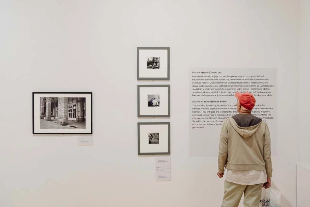 Individual wearing a red beanie and beige jacket views a wall adorned with black-and-white photographs of various sizes in an art gallery, accompanied by descriptive texts next to the artworks.