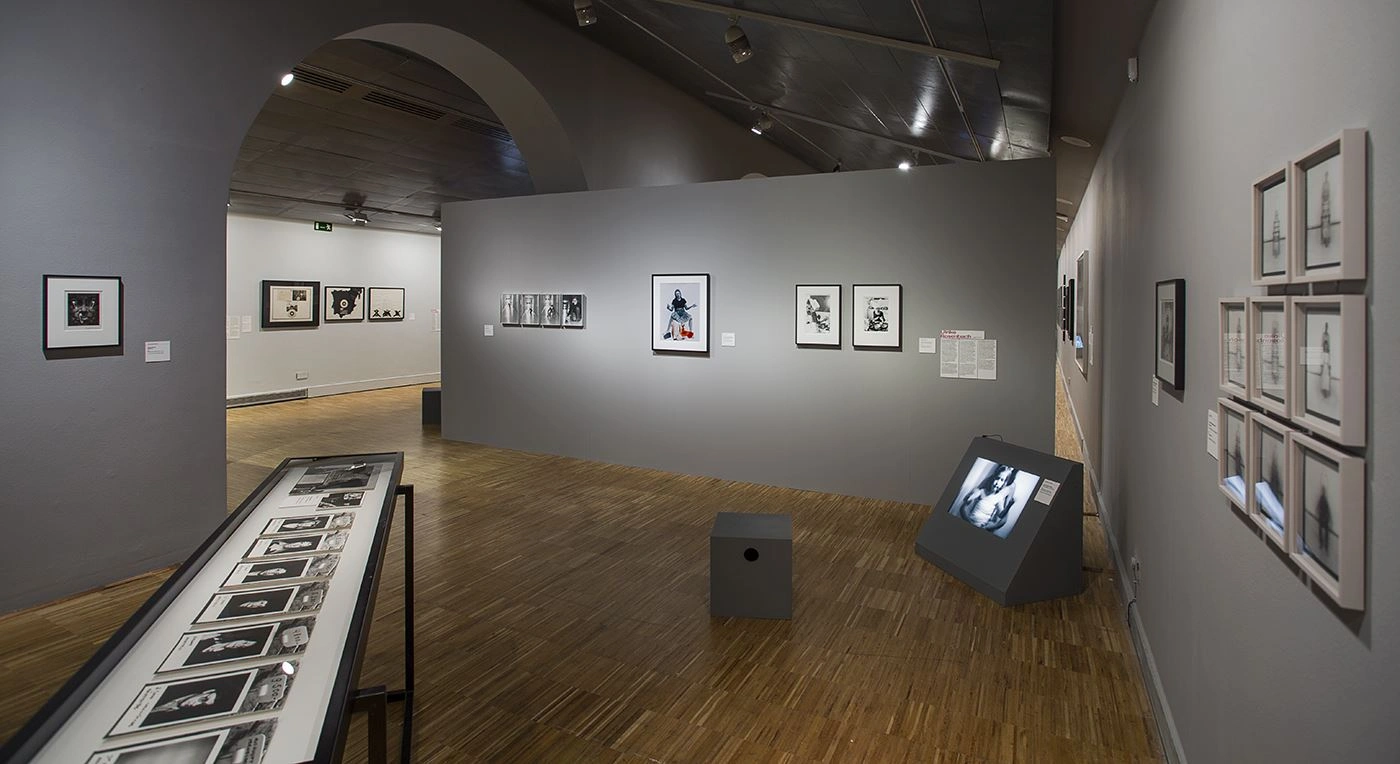 Art gallery with wooden flooring featuring multiple walls adorned with an array of framed black-and-white photographs, along with a digital display on a pedestal in the center of the room, inside a traditionally architectural space with a vaulted ceiling.