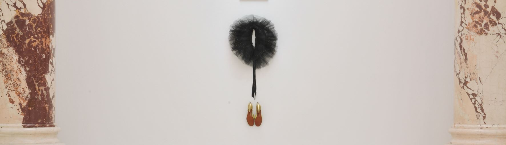 Modern art installation on a wall featuring a circular black feather object at the top, a black rod attached underneath transitioning into two golden, flame-textured shoes, against a background with an abstract brown pattern on the sides.